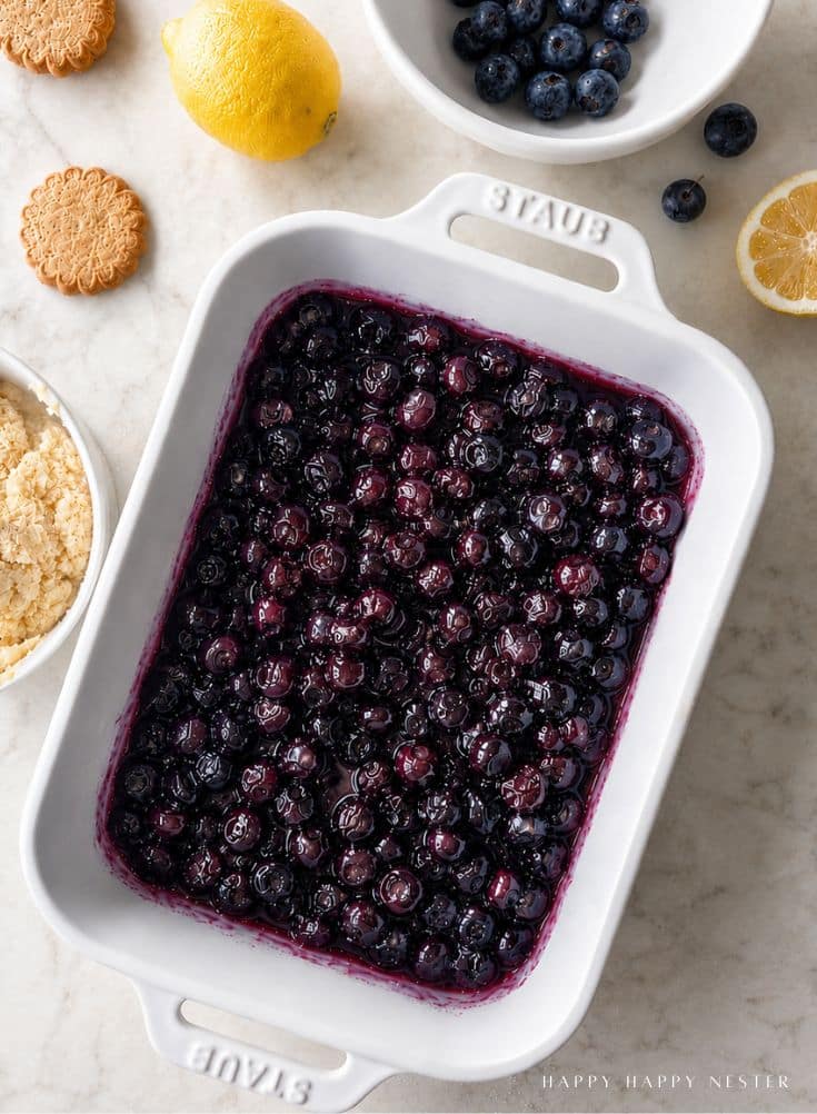 A white baking dish filled with baked blueberries sits on a marble counter, surrounded by fresh blueberries, a lemon, cookies, and crumbs in bowls.