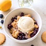 A bowl of blueberry crumble topped with a scoop of vanilla ice cream, surrounded by blueberries, lemon, cookies, and a vase with white flowers on a light surface.