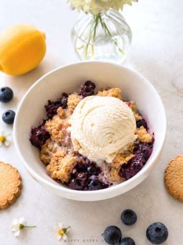A bowl of blueberry crumble topped with a scoop of vanilla ice cream, surrounded by blueberries, lemon, cookies, and a vase with white flowers on a light surface.