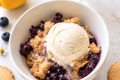 A bowl of blueberry crumble topped with a scoop of vanilla ice cream, surrounded by blueberries, lemon, cookies, and a vase with white flowers on a light surface.