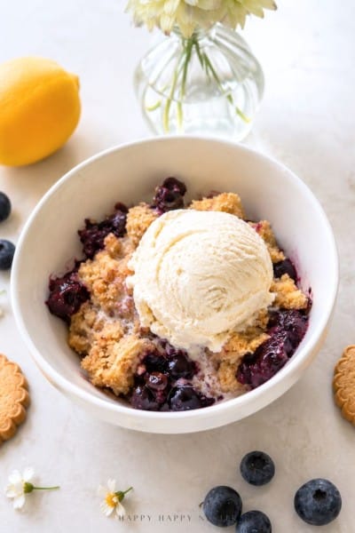 A bowl of blueberry crumble topped with a scoop of vanilla ice cream, surrounded by blueberries, lemon, cookies, and a vase with white flowers on a light surface.