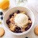 A bowl of blueberry crumble topped with a scoop of vanilla ice cream, surrounded by blueberries, lemon, cookies, and a vase with white flowers on a light surface.