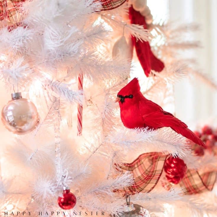 A bright red cardinal ornament sits on a white Christmas tree decorated with plaid ribbon, red and silver ornaments, and a candy cane. Soft lights illuminate the festive scene.