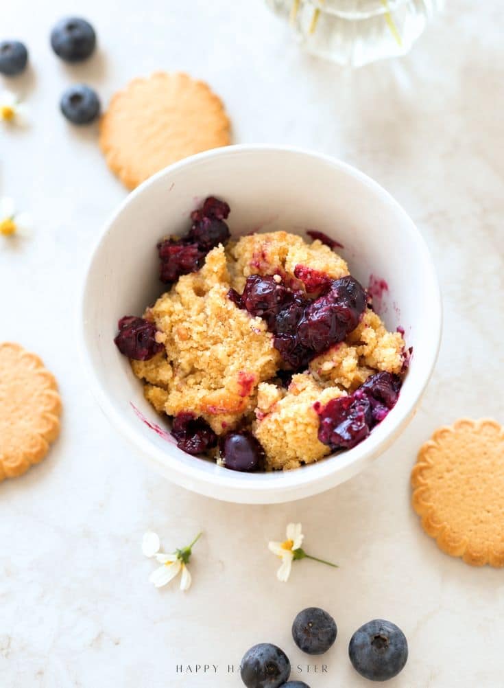 A white bowl filled with blueberry crumble sits on a light surface, surrounded by round cookies, scattered blueberries, and small white flowers.