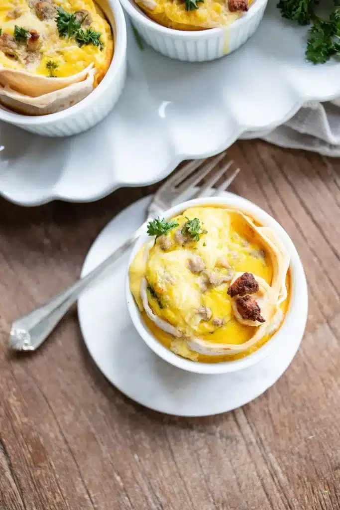 A small white ramekin filled with a baked egg, cheese, and sausage dish, garnished with parsley, sits on a white plate next to a silver fork on a wooden table.