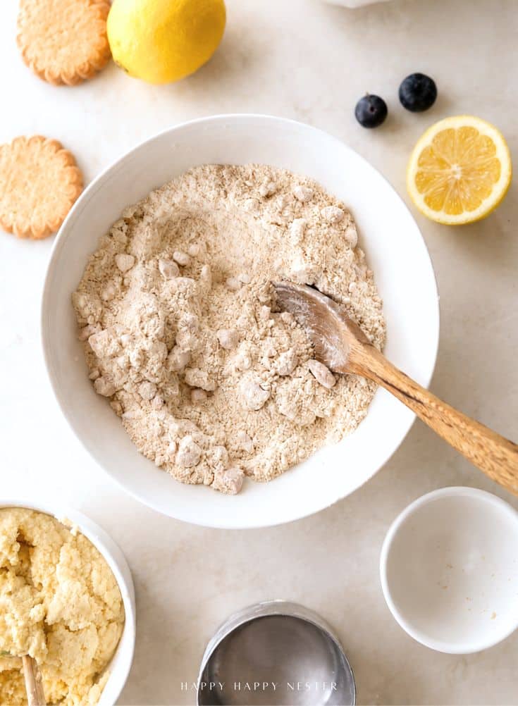 A bowl of flour mixture with a wooden spoon, surrounded by lemon halves, blueberries, cookies, a bowl of dough, and an empty white bowl on a light surface.