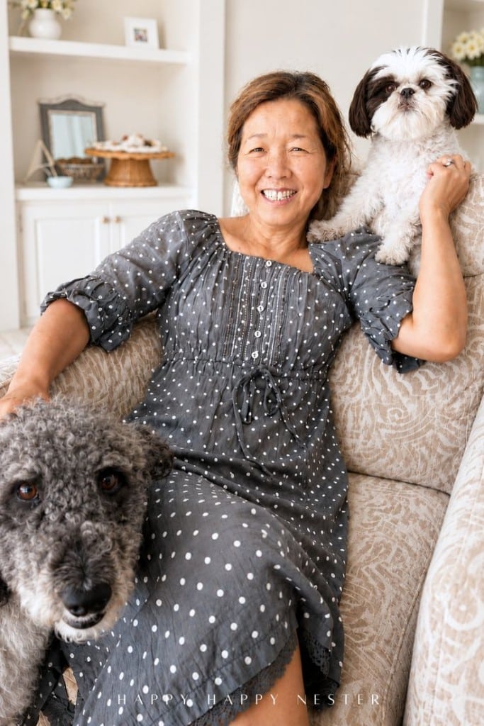 Smiling woman in a gray polka dot dress sits on a couch, holding a small white and brown dog, with a larger curly gray dog beside her. Cozy, well-lit living room background.