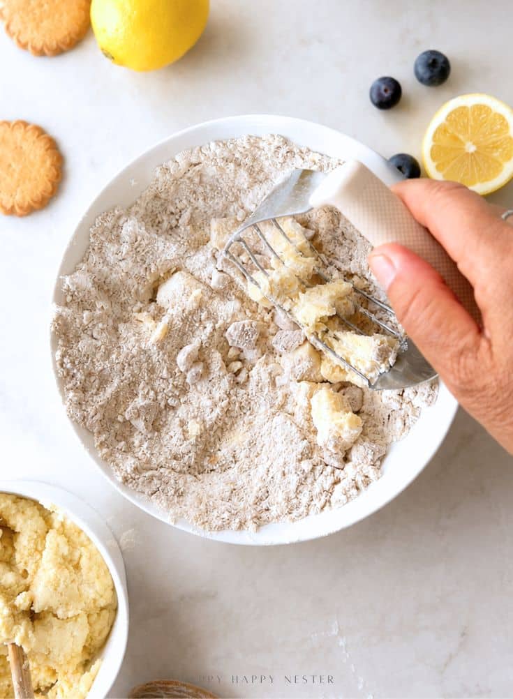 A hand uses a pastry cutter to mix dough in a white bowl on a light surface, with biscuits, blueberries, and lemon slices nearby.