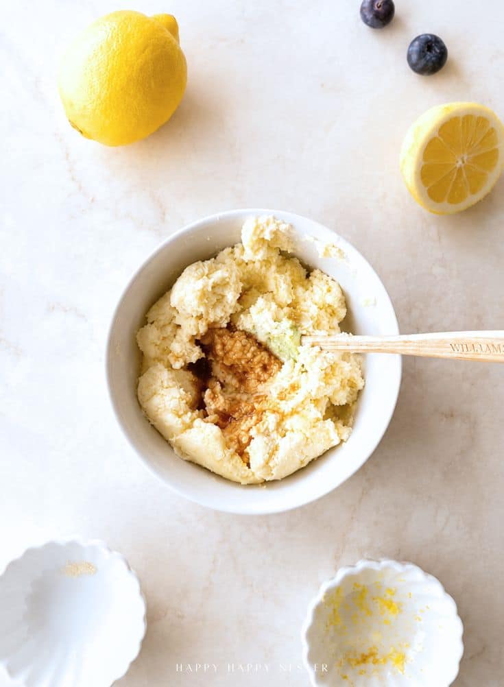 A bowl of batter with vanilla extract, surrounded by lemons, small white dishes, and a few blueberries on a light surface.