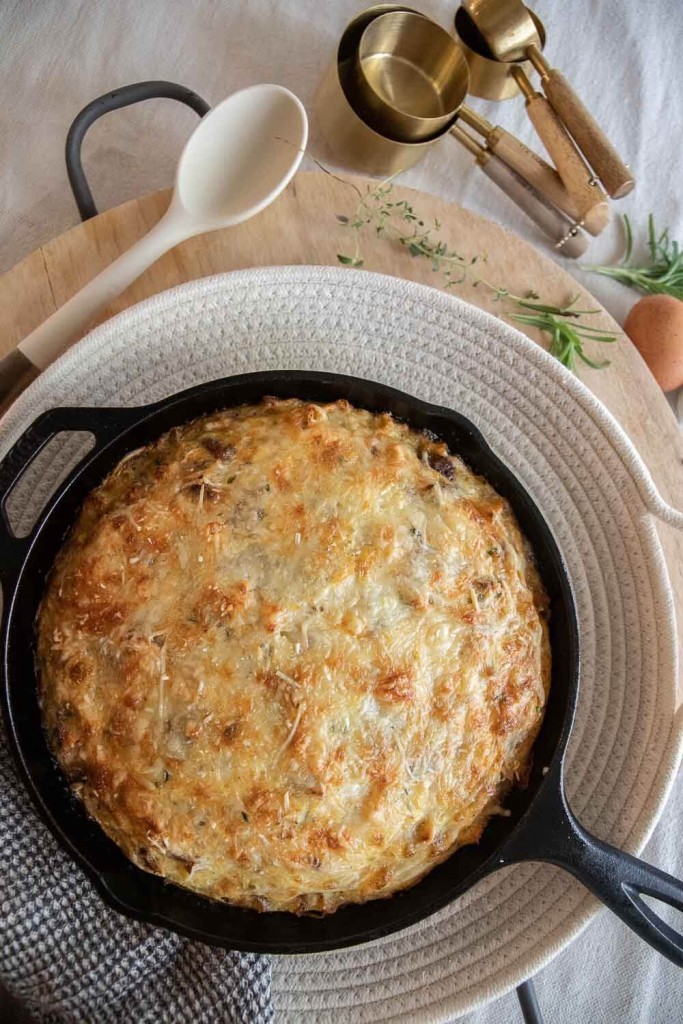 A golden, cheesy potato dish baked in a black cast iron skillet sits on a round placemat, with a wooden spoon, metal measuring cups, and fresh herbs nearby.