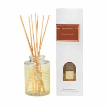 A glass diffuser bottle with wooden sticks next to a white box labeled “Cinnamon Chai.” The box has a small window showing the bottle inside.
