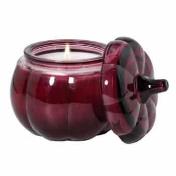 A lit white candle in a round, dark red glass jar shaped like a pumpkin, with the matching lid resting on its side.