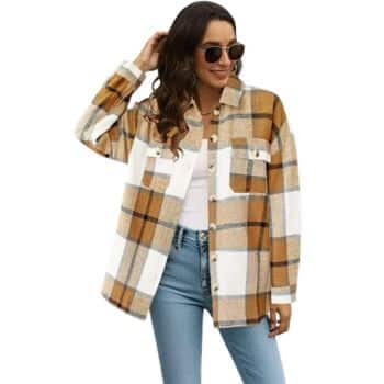 Woman wearing sunglasses, a white top, light blue jeans, and a brown, white, and beige plaid button-up shirt jacket, posing with one hand in her hair against a plain background.
