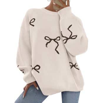 Person wearing a loose, cream-colored sweater with black bow designs, paired with light blue jeans. The person is posing with one hand on their hip and the other hand near their face, holding a phone.