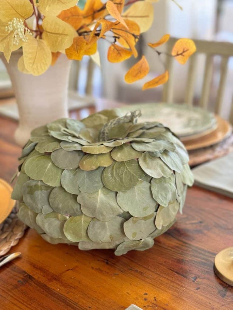A centerpiece shaped like a pumpkin, covered in green leaves, sits on a wooden table with fall-themed decor and yellow-orange foliage in the background—perfect inspiration for your next Saturday chitchat about fall crafts or swapping cozy fall recipes.