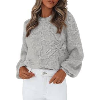 Woman wearing a light gray knit sweater with a large, textured bow design on the front, paired with high-waisted white pants. She has long, wavy hair and is posing with one hand in her pocket.