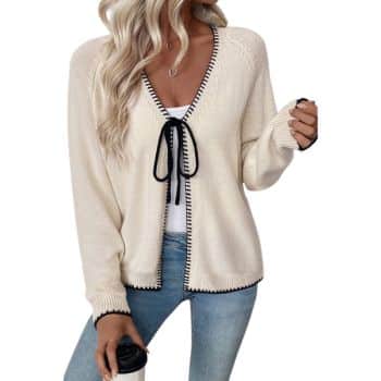 Woman wearing a cream cardigan with black trim and a bow, white top, and light blue jeans, holding a cup. Her long blonde hair is styled in loose waves.