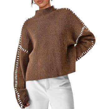 A person wearing a loose brown knit sweater with white stitching details along the sleeves and sides, paired with white pants, poses with one hand in their pocket.