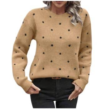 A person wearing a tan sweater with black polka dots and dark pants, standing with hands in pockets against a white background.