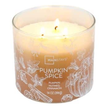 A glass jar candle labeled “Pumpkin Spice” with three lit wicks. The jar is decorated with white illustrations of pumpkins, cinnamon sticks, and nutmeg. The candle wax is light orange.