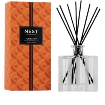 A Nest Fragrances Pumpkin Chai reed diffuser with a geometric orange box on the left and a clear glass bottle with black reeds on the right.