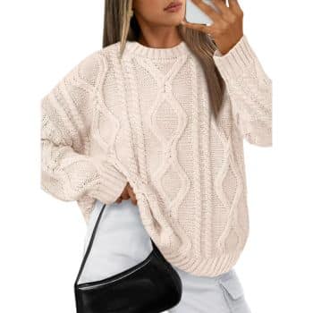 Woman wearing a loose, cream cable knit sweater, white pants, and holding a small black shoulder bag, taking a mirror selfie with her phone.