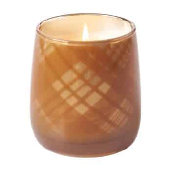 A lit candle in a brown glass holder with a plaid pattern, casting a warm, soft glow.