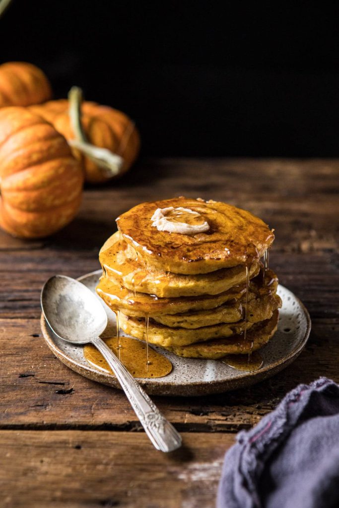 A stack of pancakes topped with butter and syrup sits on a rustic plate with a spoon beside it. In the background, pumpkins rest on a wooden table, setting the perfect scene for Saturday chitchat and cozy fall recipes.