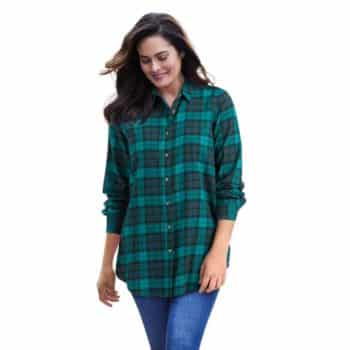 A woman with long brown hair wears a green and black plaid button-up shirt with blue jeans, standing and smiling against a white background.