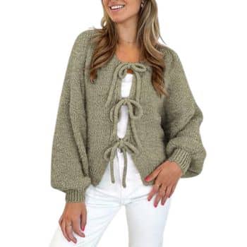 A woman wears a sage green, chunky-knit cardigan with long balloon sleeves and three ribbon ties down the front, worn over a white top and white pants. The background is plain white.