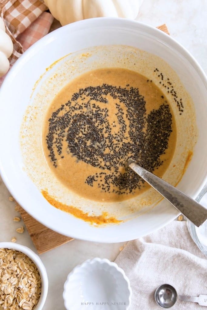 A mixing bowl with pumpkin batter and chia seeds being stirred with a spoon. Oats, a napkin, and a small white dish are nearby on the counter.