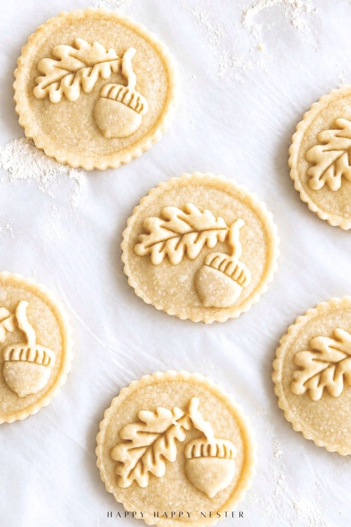 Round cookie dough with scalloped edges, each decorated with a detailed oak leaf and acorn design, arranged on a floured white surface ready for baking.