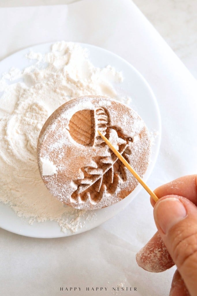 A hand uses a toothpick to remove flour from a wooden cookie stamp with an acorn and oak leaf design, perfect for shaping shortbread cookies. Flour is scattered on a white plate in the background.
