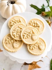 Six round cookies with embossed oak leaves and acorns are arranged on a white cake stand. A small white pumpkin and autumn leaves are in the background, suggesting a fall or Thanksgiving theme.