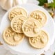 Six round cookies with embossed oak leaves and acorns are arranged on a white cake stand. A small white pumpkin and autumn leaves are in the background, suggesting a fall or Thanksgiving theme.