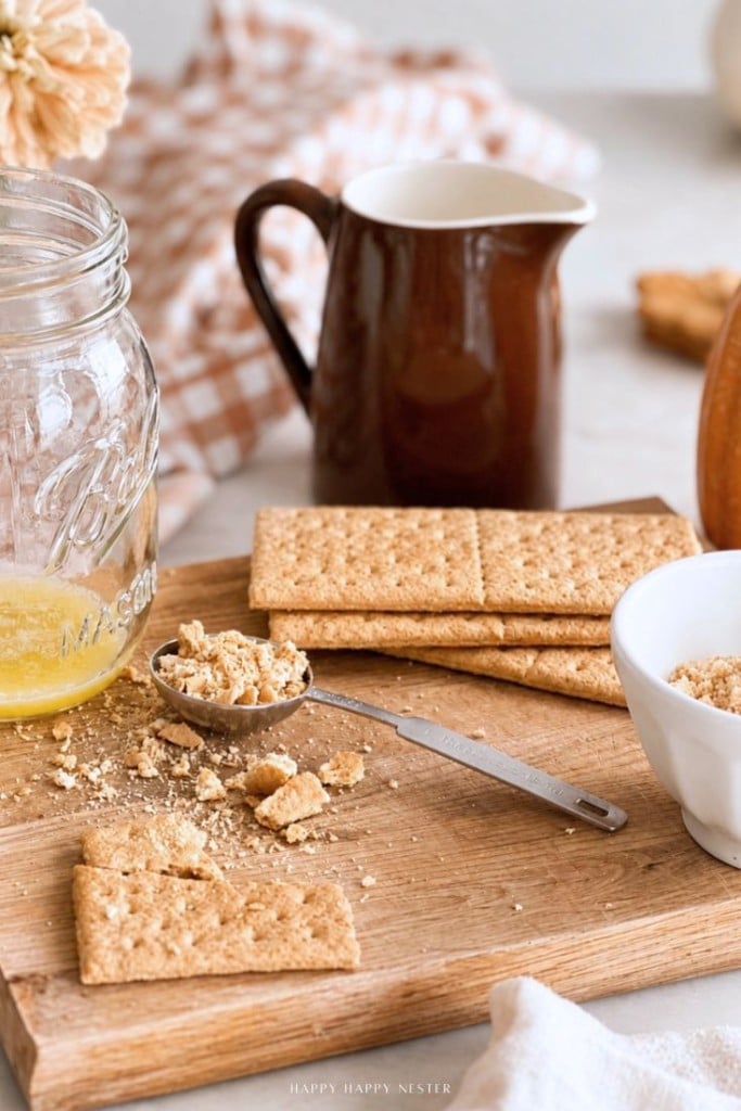 A wooden board with graham crackers, a spoon with crumbs, a glass jar with melted butter, a small white bowl, and a brown pitcher. A checkered cloth and a flower are in the background.