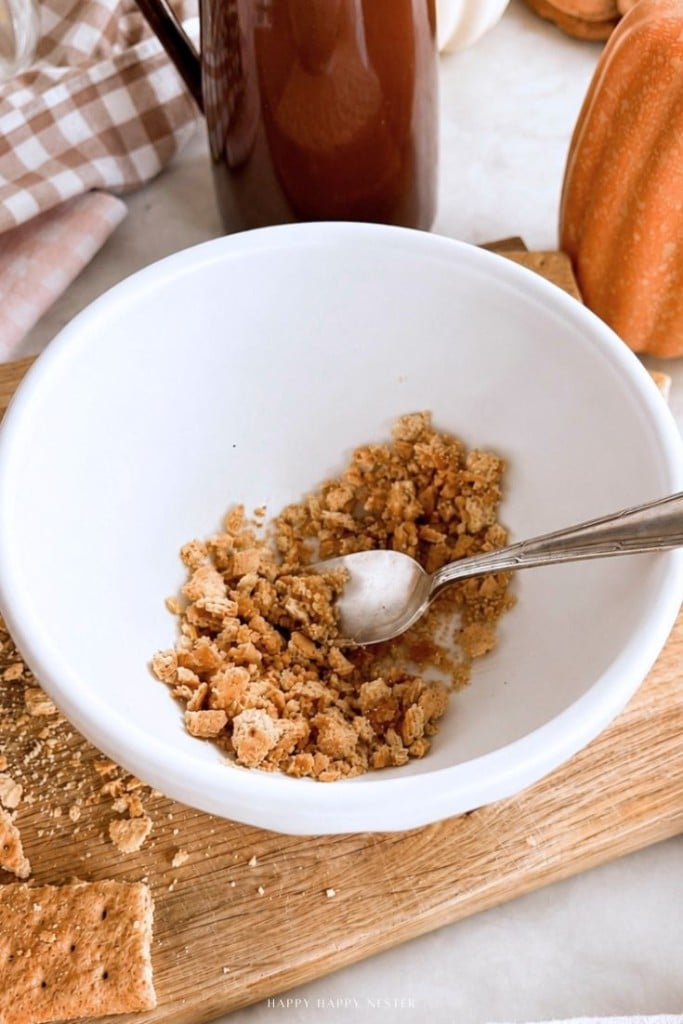 A white bowl with crushed graham crackers and a spoon sits on a wooden cutting board. Some crumbs and a graham cracker are scattered nearby. A pumpkin and a brown pitcher are in the background.