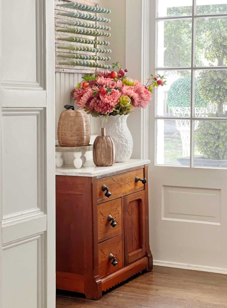 A wooden cabinet with black knobs holds a white vase of pink flowers, a woven pumpkin, and charming fall decor—setting the perfect scene for Saturday Chitchat or sharing your favorite Fall Recipes—near a window with a view of greenery outside. Light streams through the glass panes.