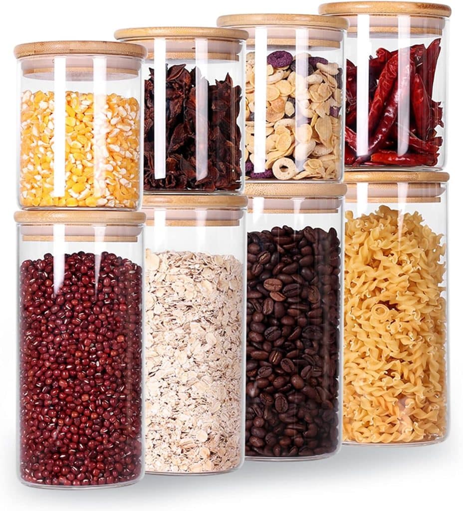 Eight clear glass jars with bamboo lids, each filled with various dry foods including corn, red beans, oats—perfect for making pumpkin overnight oats—pasta, coffee beans, chili peppers, dried fruits, and other legumes, arranged in two tidy rows.