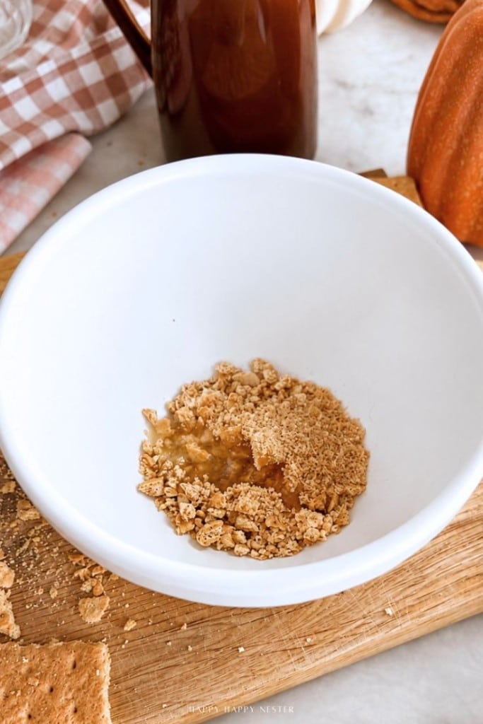 A white bowl with crushed graham crackers and melted butter sits on a wooden board, surrounded by baking ingredients and a checkered cloth.