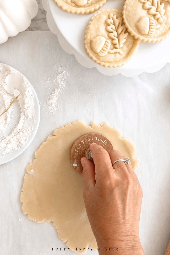 A hand presses a round cookie stamp onto rolled dough. A plate with flour and shaped, stamped cookies are nearby on a white surface.