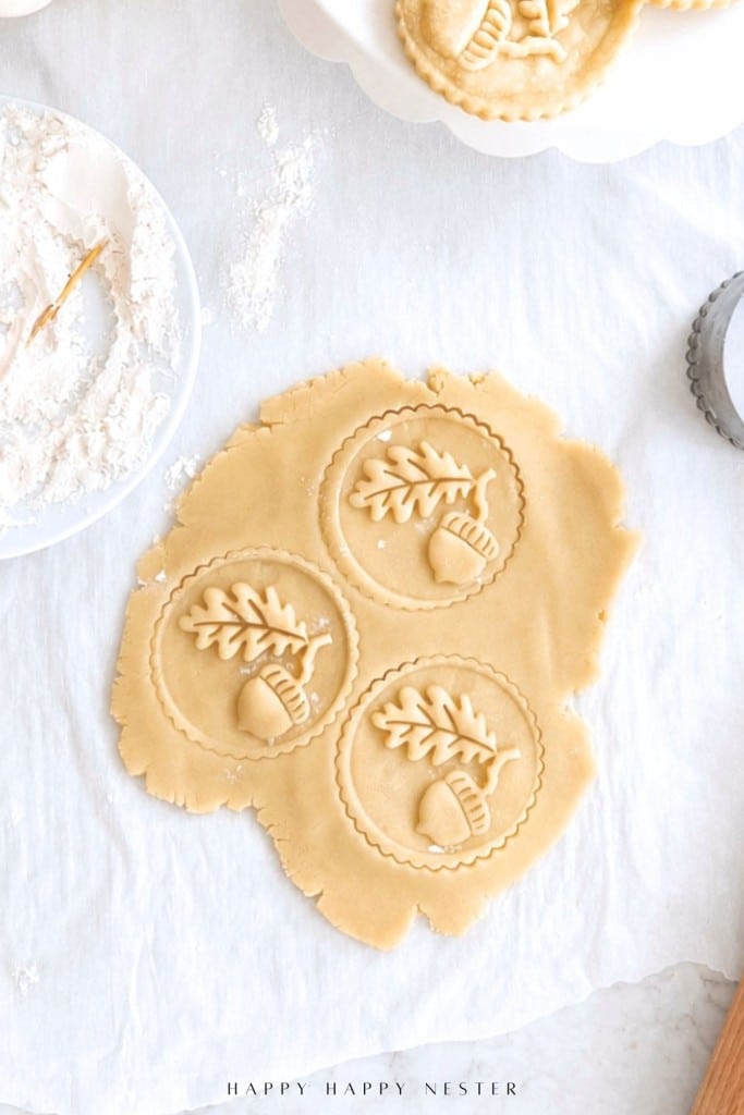 Rolled-out cookie dough with three round shapes featuring an acorn and leaf design pressed into them, surrounded by baking tools and dusted flour on a white surface.