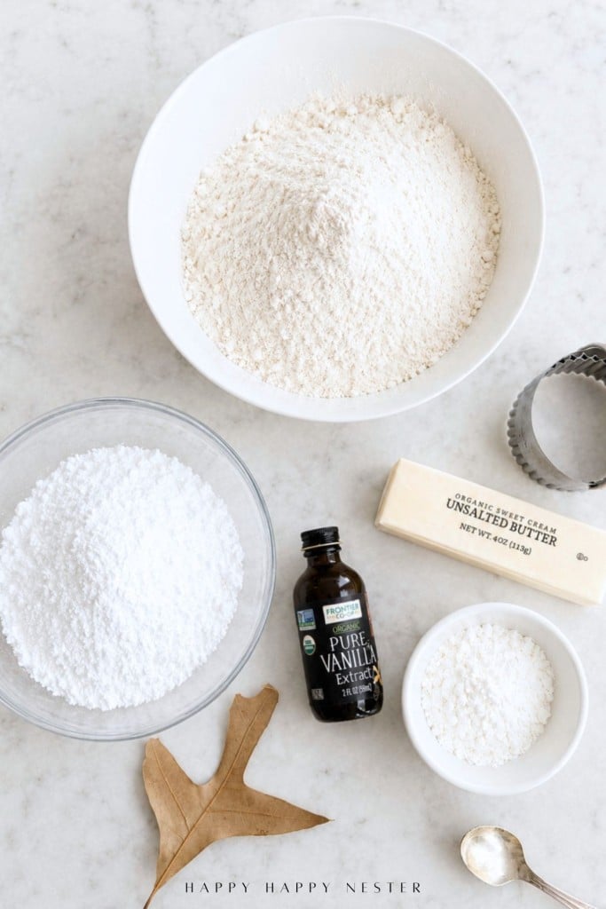 White bowls with flour and powdered sugar, a stick of unsalted butter, a small dish of sugar, a bottle of vanilla extract, a metal cookie cutter ready for cookie stamp shortbread cookies, a dried oak leaf, and a gold spoon all arranged on a light surface.
