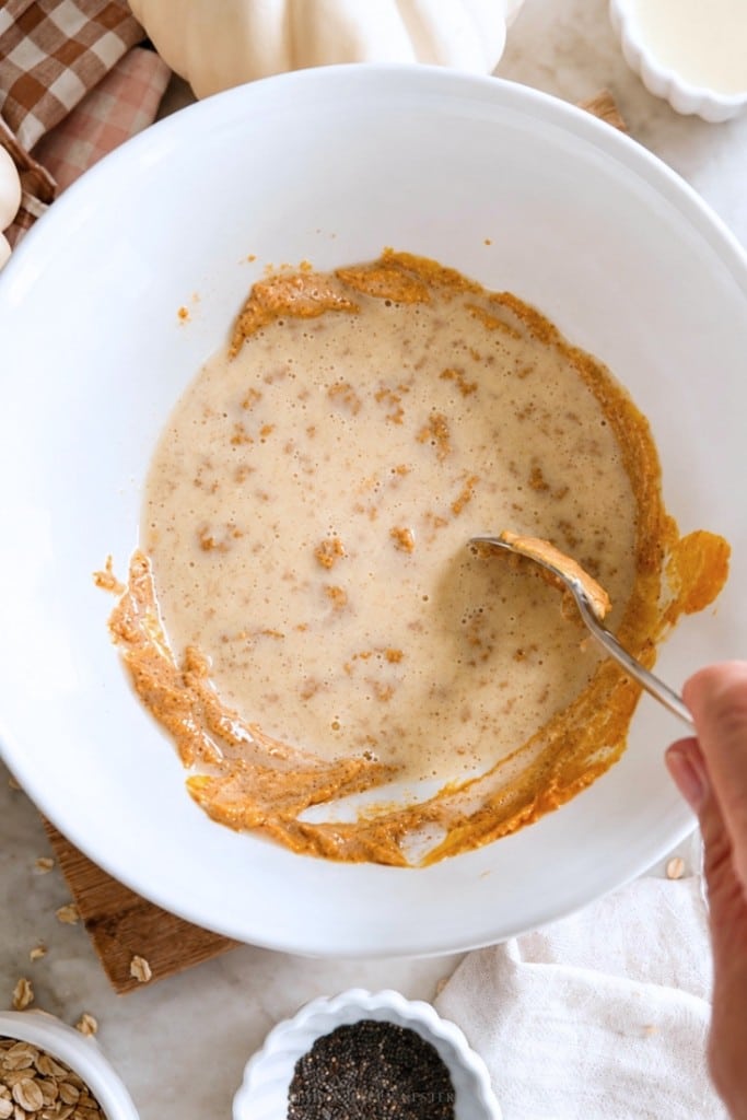 A hand stirs a mixture of wet ingredients in a white bowl with a spoon. Small bowls of oats and seeds are nearby on a marble countertop.