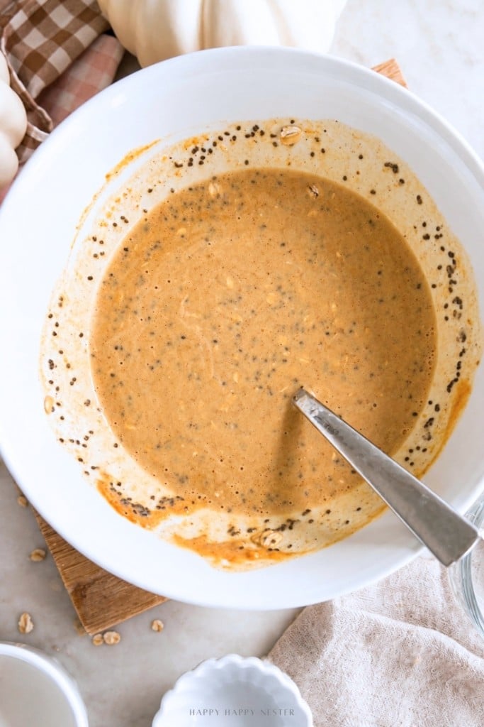 A white bowl filled with pumpkin-colored batter mixed with oats and chia seeds, with a metal spoon inside, sitting on a light countertop near a beige cloth and small pumpkins.