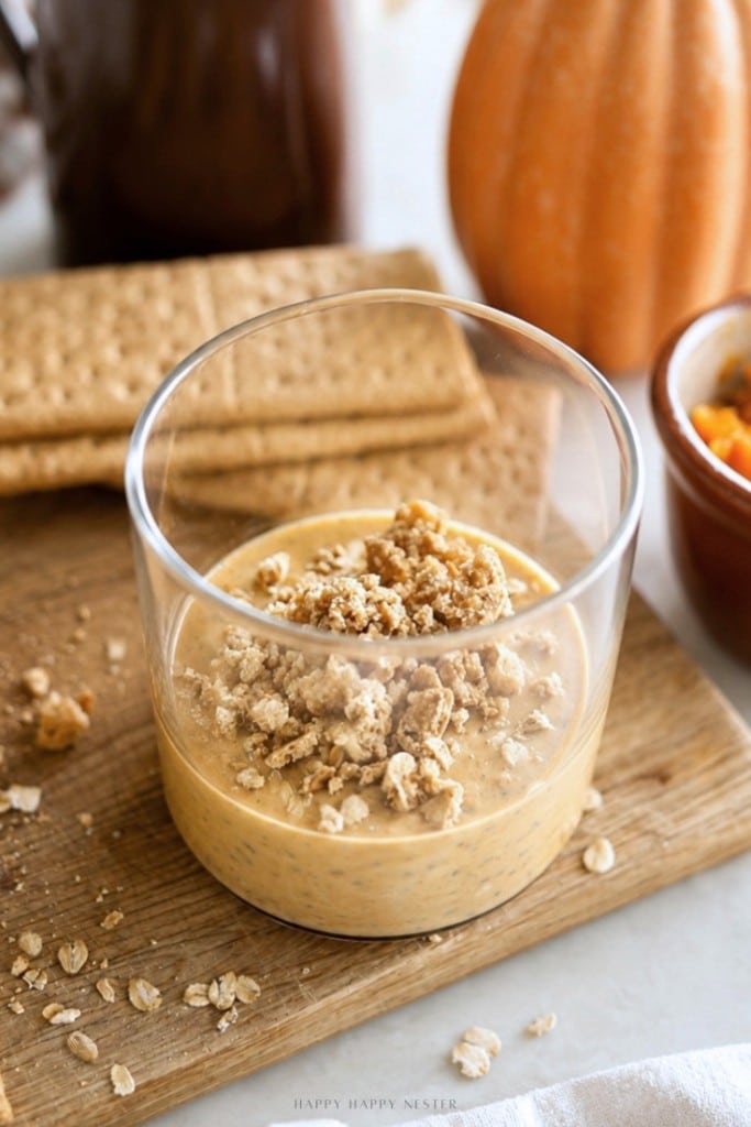 A glass cup filled with creamy pumpkin dessert, reminiscent of pumpkin overnight oats, is topped with oat crumble and sits on a wooden board with graham crackers, oats, and a small pumpkin nearby.