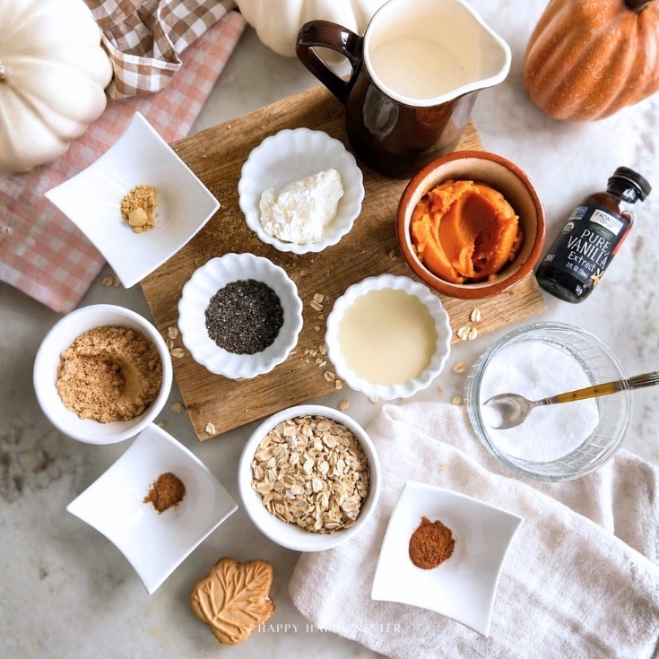 A flat lay of pumpkin pie overnight oats ingredients in bowls, including oats, pumpkin puree, brown sugar, spices, chia seeds, milk, vanilla, and a pitcher, on a wooden board with fall decor and a checkered napkin.