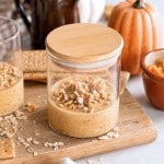 A glass jar of pumpkin parfait topped with granola sits on a wooden board, surrounded by graham crackers, oats, a pumpkin, and a brown pitcher, evoking an autumn theme reminiscent of cozy mornings with pumpkin overnight oats.