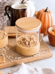 A glass jar of pumpkin parfait topped with granola sits on a wooden board, surrounded by graham crackers, oats, a pumpkin, and a brown pitcher, evoking an autumn theme reminiscent of cozy mornings with pumpkin overnight oats.
