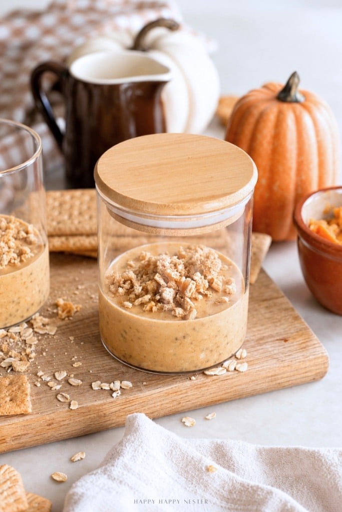 A glass jar of pumpkin parfait topped with granola sits on a wooden board, surrounded by graham crackers, oats, a pumpkin, and a brown pitcher, evoking an autumn theme reminiscent of cozy mornings with pumpkin overnight oats.